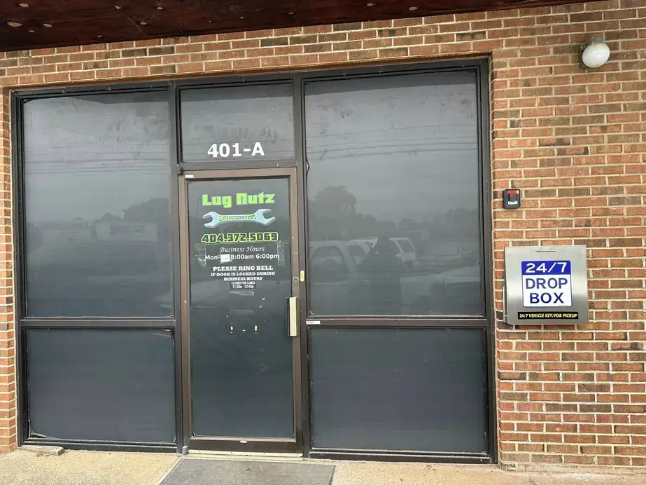 The entrance to Lug Nutz Automotive at 401-A Old Mill Road, showing the door decal with the wordmark, phone number and business hours, and the 24/7 key drop box mounted on the brick beside it